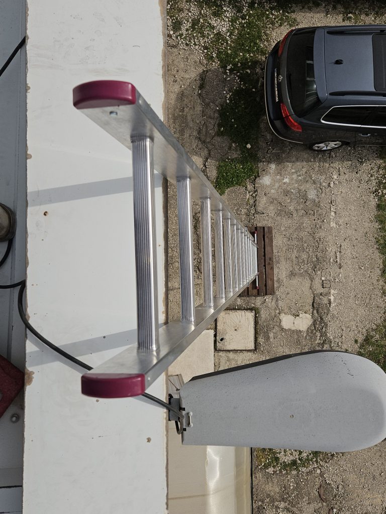 Anlegeleiter am Dachrand einer Industriehalle; Blick nach unten auf Hof und geparkten Pkw – sicherer Zugang für Dachwartung und Reparaturarbeiten.