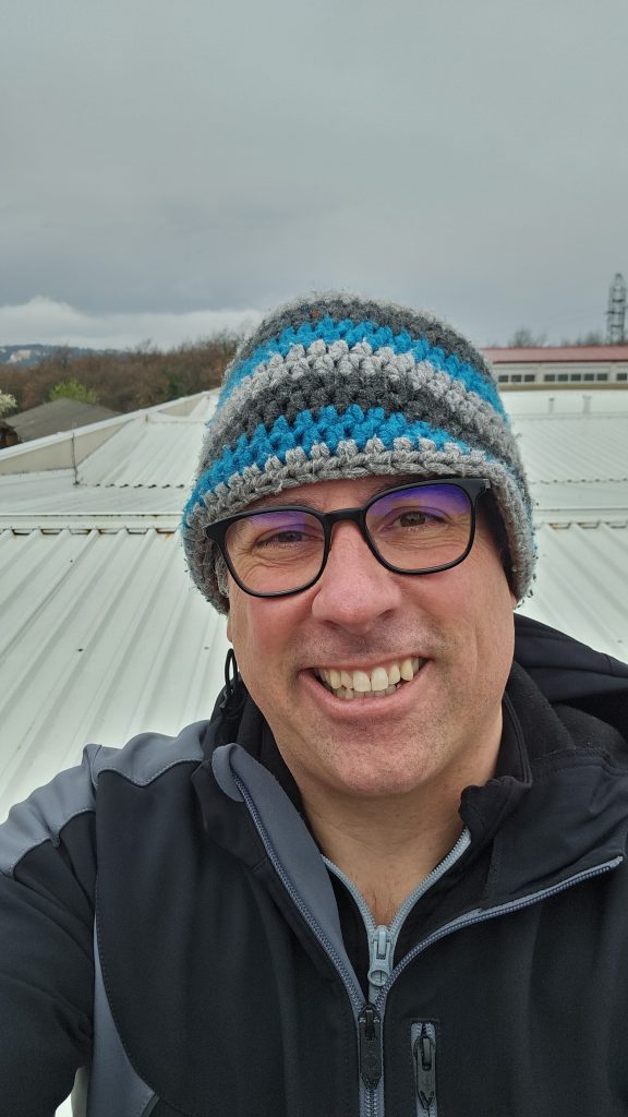 Techniker führt Wartungs- und Abdichtungsarbeiten am Trapezblechdach einer Halle aus; Kontrolle des Attika-/Rinnenanschlusses neben Kaminwand